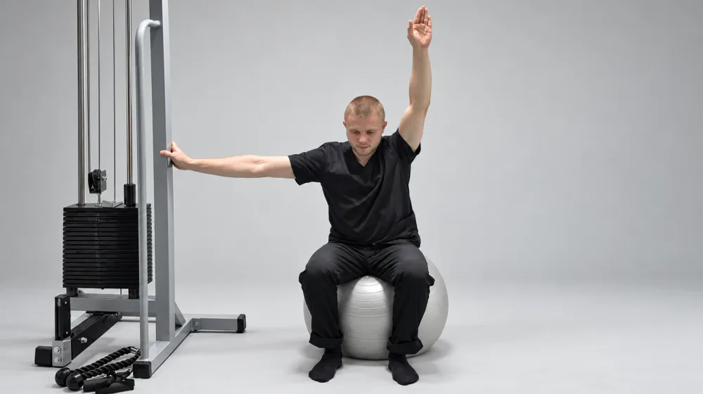 Корригирующее упражнение при сколиозе: сидя на фитболе, одна рука на опоре, другая вытянута вверх